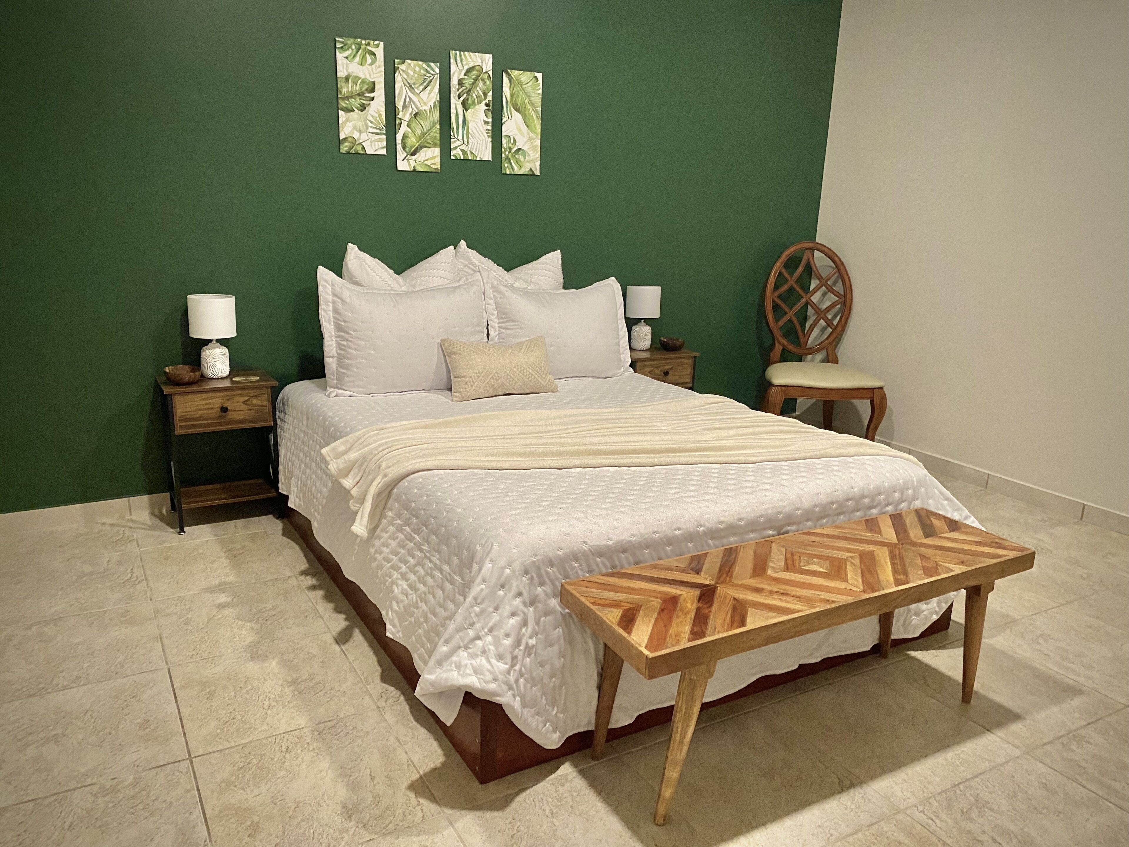 Une chambre à coucher comprenant un lit, deux tables de chevet, un banc en bois, une chaise et un mur orné de quatre tableaux encadrés.