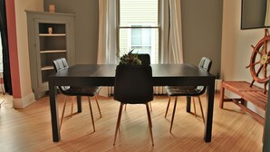 Un comedor con una mesa y sillas negras, piso de madera, una ventana con cortinas, una estantería y un adorno de timón.