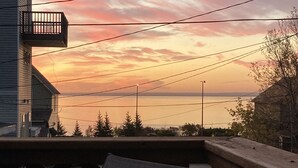 Un balcón con vista a un atardecer sobre un cuerpo de agua, líneas eléctricas y un edificio también con balcón.