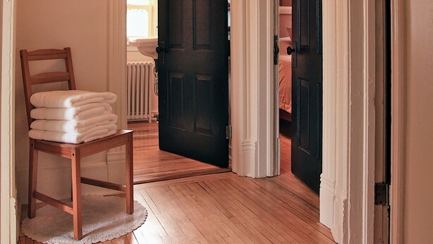 Un pasillo con suelo de madera, una silla con toallas dobladas y una puerta que da a otra habitación.