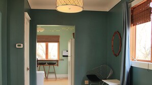 Una habitación con paredes verde azulado, suelo de madera, alfombra estampada, una silla, una mesa, un radiador y una ventana con cortinas.