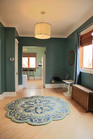 Una stanza con pareti color acquamarina, pavimento in legno, tappeto decorato, una sedia, un tavolo, un termosifone e una finestra con tende.