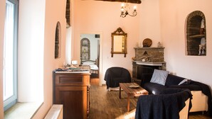 Een gezellige woonkamer met een open haard, houten balken in het plafond, een spiegel en een zicht op een slaapkamer door een boog.