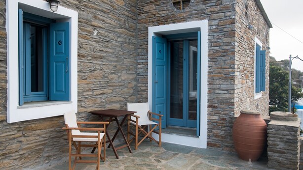 Een stenen gebouw met blauwe deuren en ramen, een houten tafel met stoelen en een grote terracotta pot.