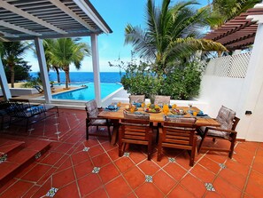 Eine Terrasse mit einem Holztisch, gedeckt für das Frühstück, mit Blick auf einen Pool und das Meer.