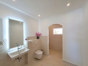 Ein modernes Badezimmer mit Waschbecken, Spiegel und Toilette in Weiß. Ein kleines Fenster bietet Blick auf Grünflächen.