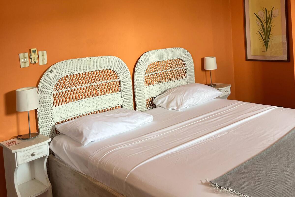 Una habitación con pared naranja, una cama con ropa de cama blanca, dos mesitas de noche con lámparas y un ventilador de techo.