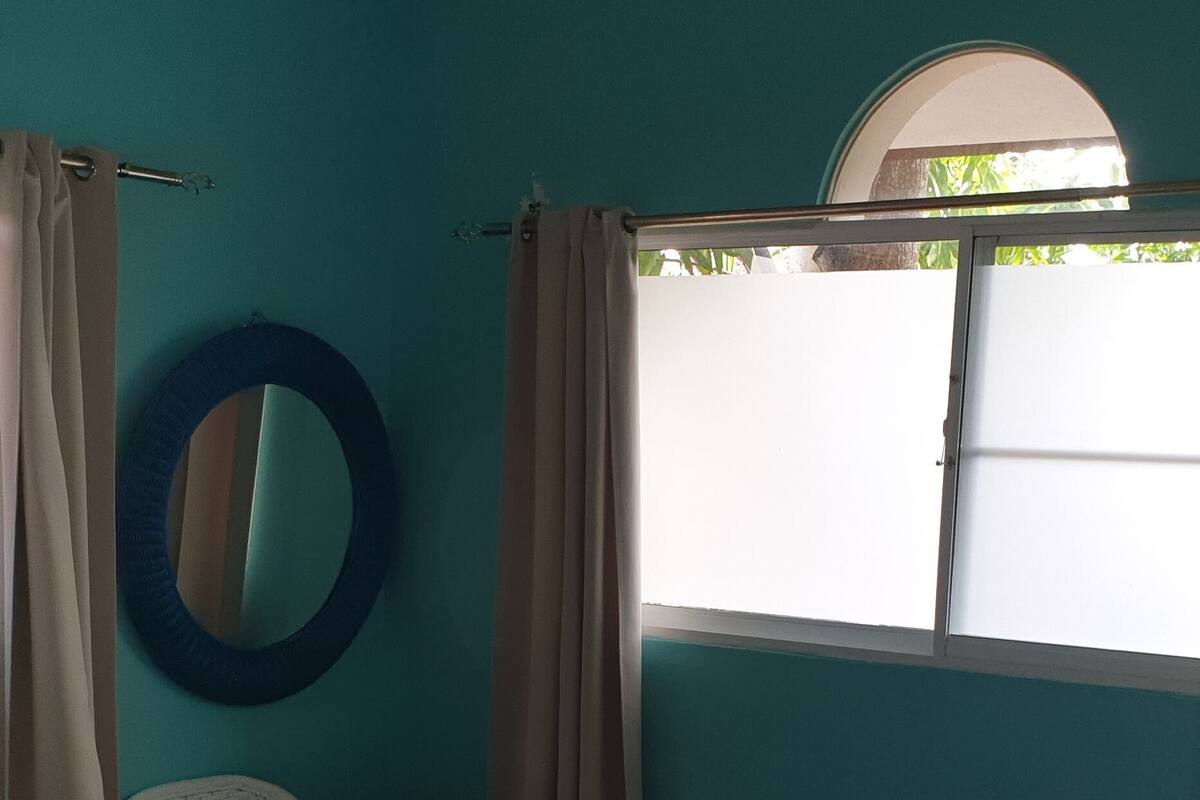 Un dormitorio con cama, ventilador de techo, ventana con cortinas y un espejo montado en la pared.