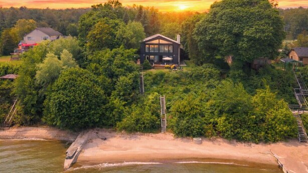 A modern house with a large deck, surrounded by dense trees and a lake. - Blue Water Beach House! Amazing Beach, sleeps 20! (Sanilac Township)