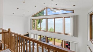A spacious room with large windows overlooking a body of water, a ceiling fan, and a wooden railing. - Blue Water Beach House! Amazing Beach, sleeps 20! (Sanilac Township)
