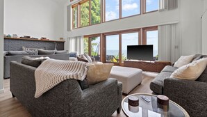 A spacious living room with a high ceiling, large windows offering a view of the outdoors, a flat-screen TV, and a cozy seating area. - Blue Water Beach House! Amazing Beach, sleeps 20! (Sanilac Township)