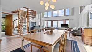 A spacious living area with a wooden staircase, a dining table, and a large window offering a view of the outdoors. - Blue Water Beach House! Amazing Beach, sleeps 20! (Sanilac Township)