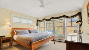 A bedroom with a wooden bed, two bedside tables, a desk, and a window with a view of autumn foliage.