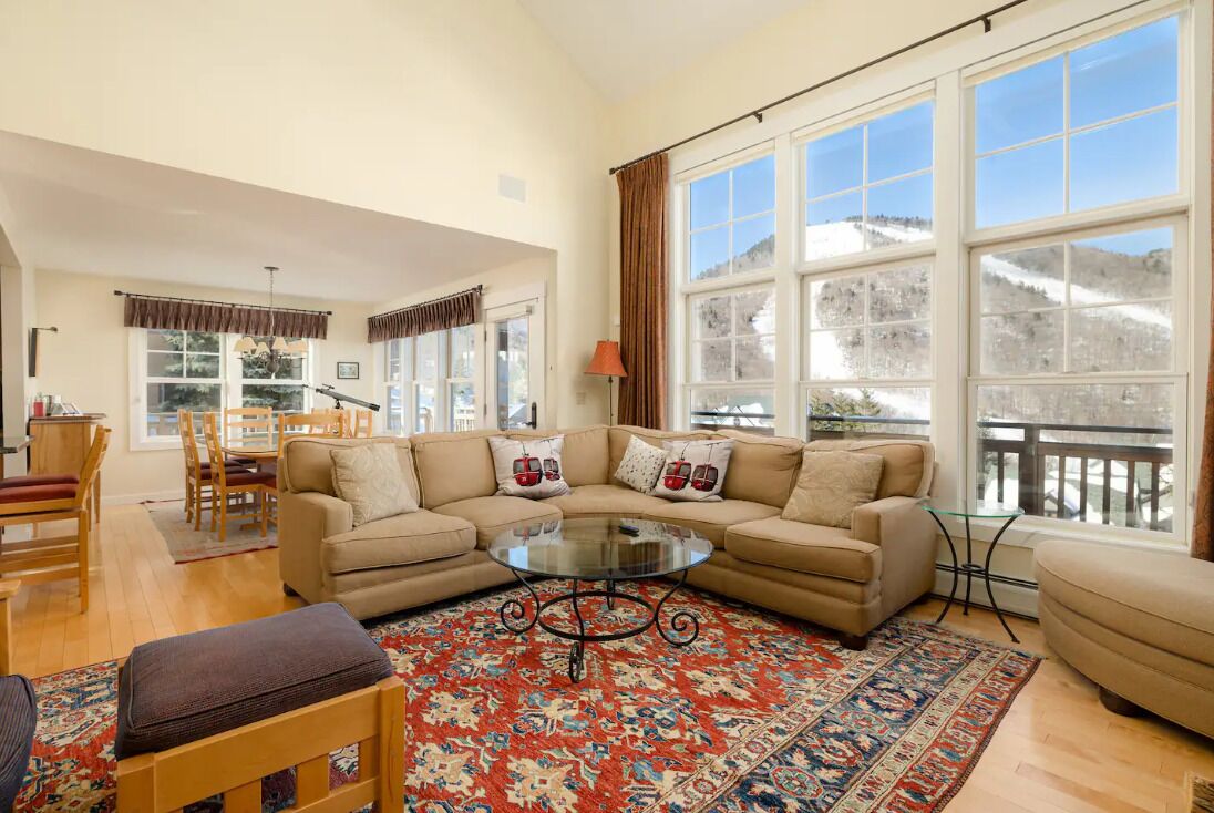 A living room with a beige sofa, a glass coffee table, and a patterned rug. There are large windows with curtains, a dining area with wooden furniture, and a view of a snowy landscape.