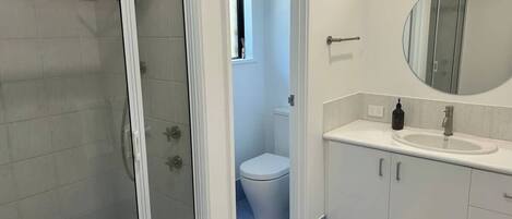 Baño con cabina de ducha de vidrio, inodoro, lavamanos con espejo y un ventanal pequeño.