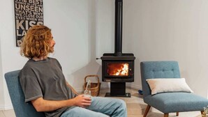 Una persona sentada en una silla en una habitación con un estufa de leña, un sillón azul y una cesta de mimbre.