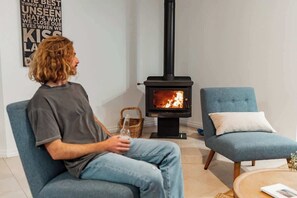 A person sitting on a chair in a room with a wood-burning stove, a blue armchair, and a wicker basket.