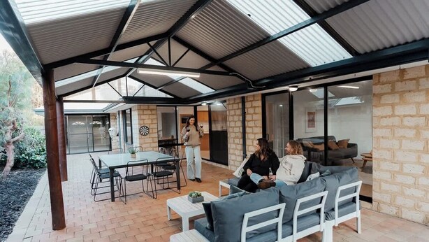 Un patio exterior con una zona de estar cubierta, una mesa de comedor y una cocina visible a través de puertas de cristal.