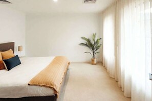 A bedroom with a bed, pillows, a bedside table, a lamp, a plant, and sheer curtains.