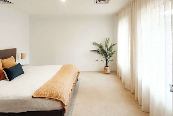 A bedroom with a bed, pillows, a bedside table, a lamp, a plant, and sheer curtains.