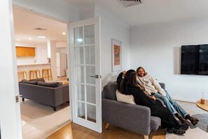 A modern living room with a grey sofa, a dining area with wooden stools, and a kitchen in the background.