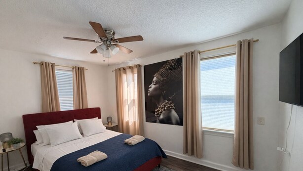 A bedroom with a bed, a ceiling fan, a TV, and a window with curtains.