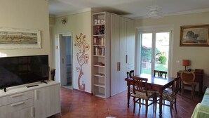 A living room with a TV, a dining table with chairs, a bookshelf, and a sliding door leading to a patio. - La Casina (Barga)