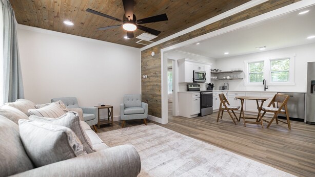 Uma sala de estar moderna com sofá, poltronas e uma mesa de jantar. Cozinha com armários brancos e um espaço de refeições é visível por uma porta aberta.