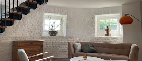 Una sala de estar con una escalera, un sofá, un sillón, una mesita y una planta en maceta.