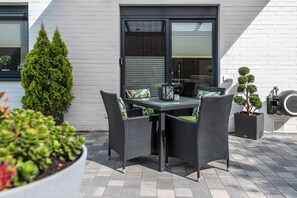Un patio al aire libre con una mesa y sillas de comedor, rodeado de plantas en macetas y una pared de ladrillo blanco.