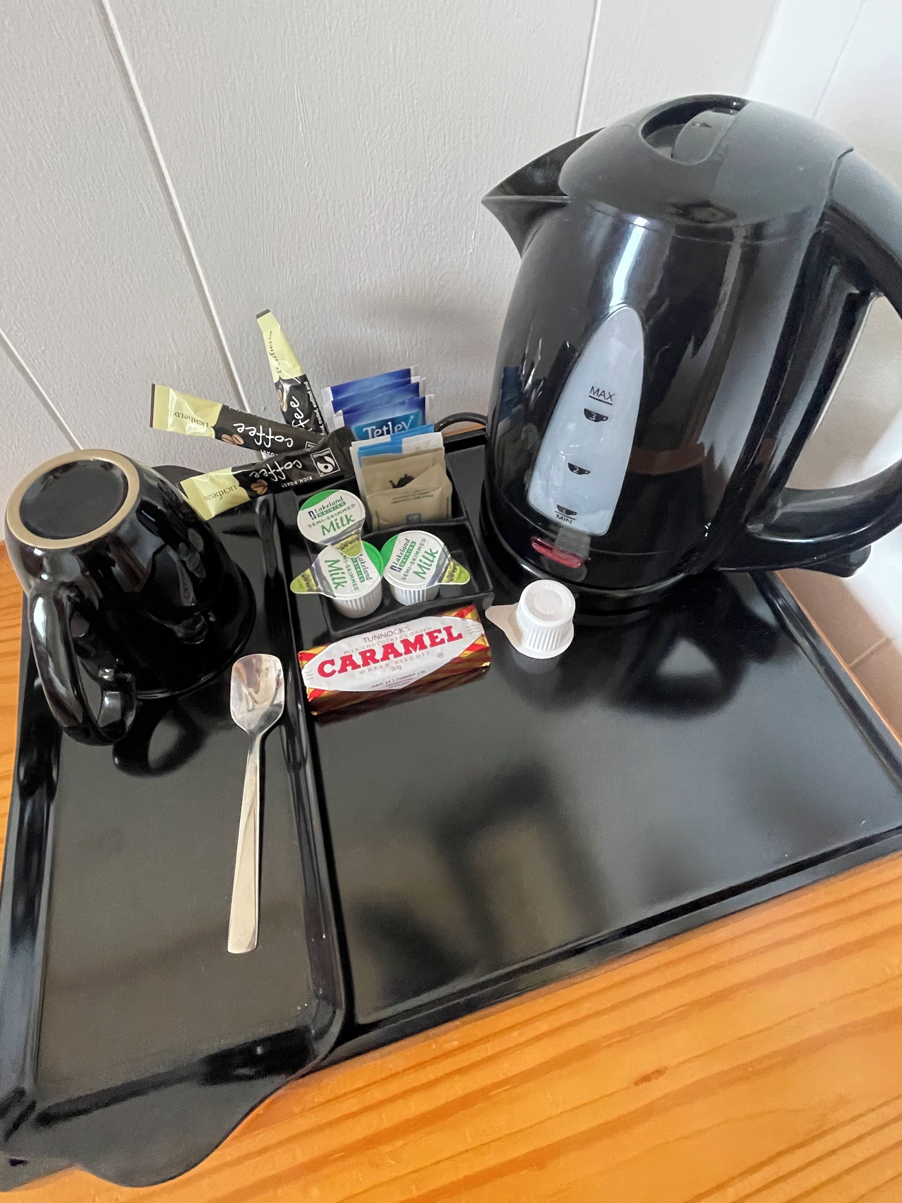 Un plateau avec une cafetière noire, une cuillère, une boîte de caramels et plusieurs sachets de lait et de crème pour café.