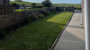A view of a grassy area with a stone wall and a paved path leading to a gate. - Broadhaven Road (Wick)