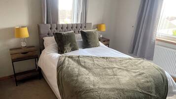 Una cama bien hecha con cabecera gris, dos almohadas y una manta verde. Hay dos mesitas de noche con lámparas a cada lado de la cama.