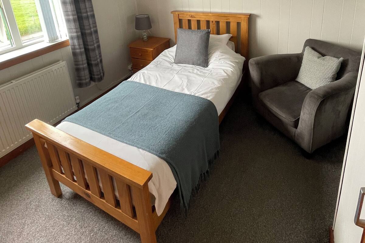 A bedroom with a bed, a chair, a nightstand, a lamp, a window with curtains, a radiator, and a ceiling light.