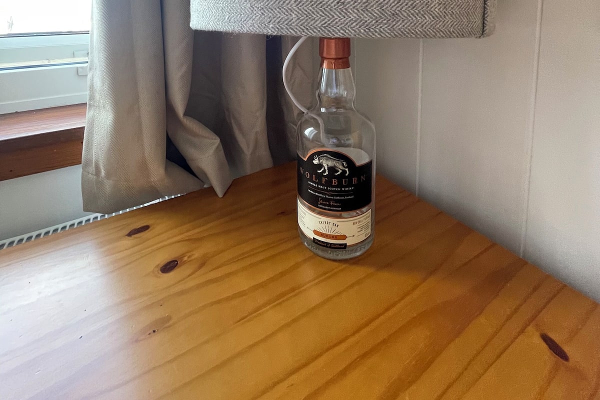 A bottle of Wolfburn Cherry Bay whisky on a wooden table with a grey textured lamp beside it.