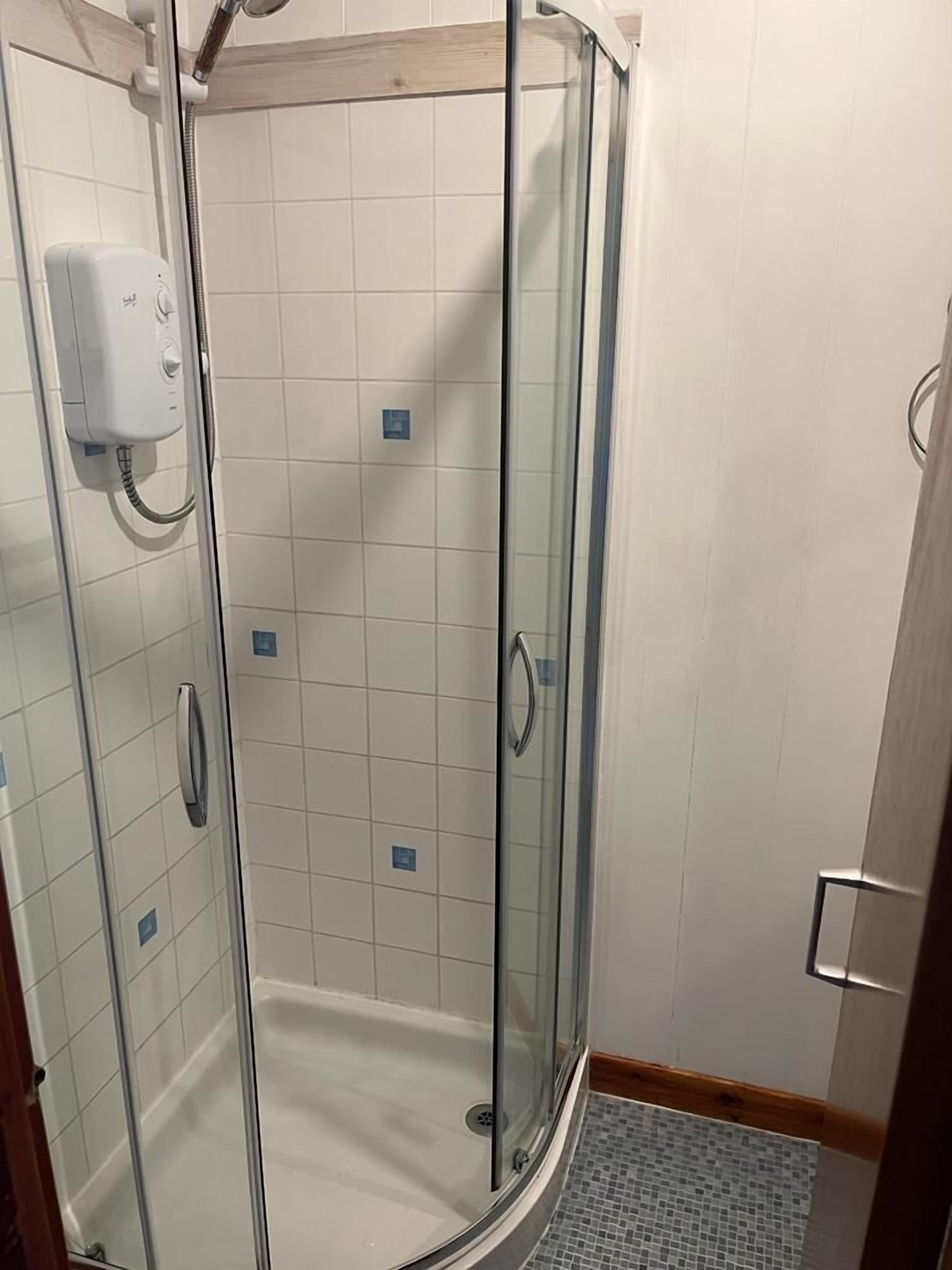 Een douchecabine met glazen wanden, een handdouche, betegelde wanden en een klein afvoerputje.