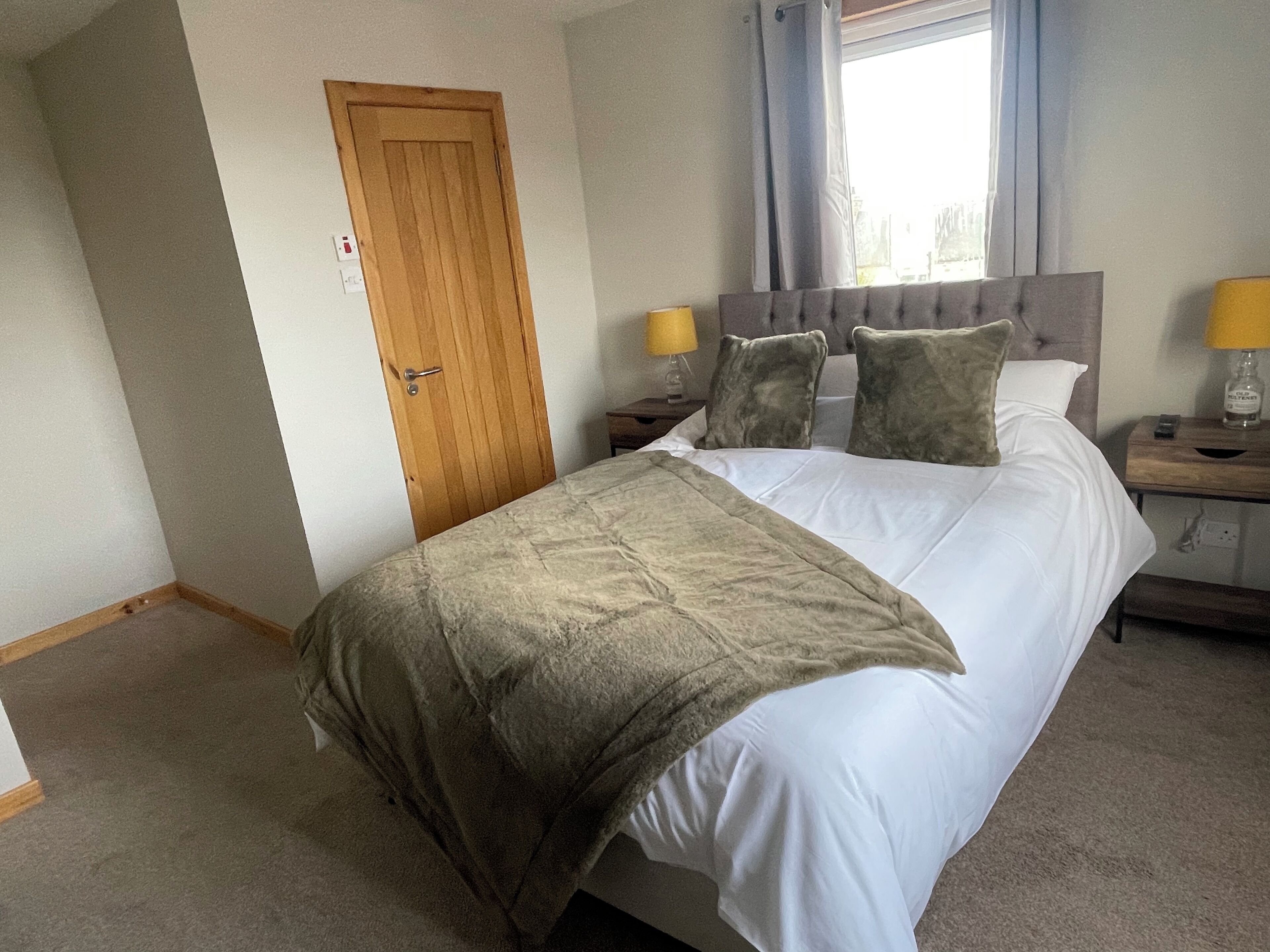 Een slaapkamer met een bed, een houten deur, een nachtkastje met een lamp en een raam met gordijnen.