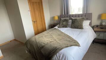 Een slaapkamer met een bed, een houten deur, een nachtkastje met een lamp en een raam met gordijnen.