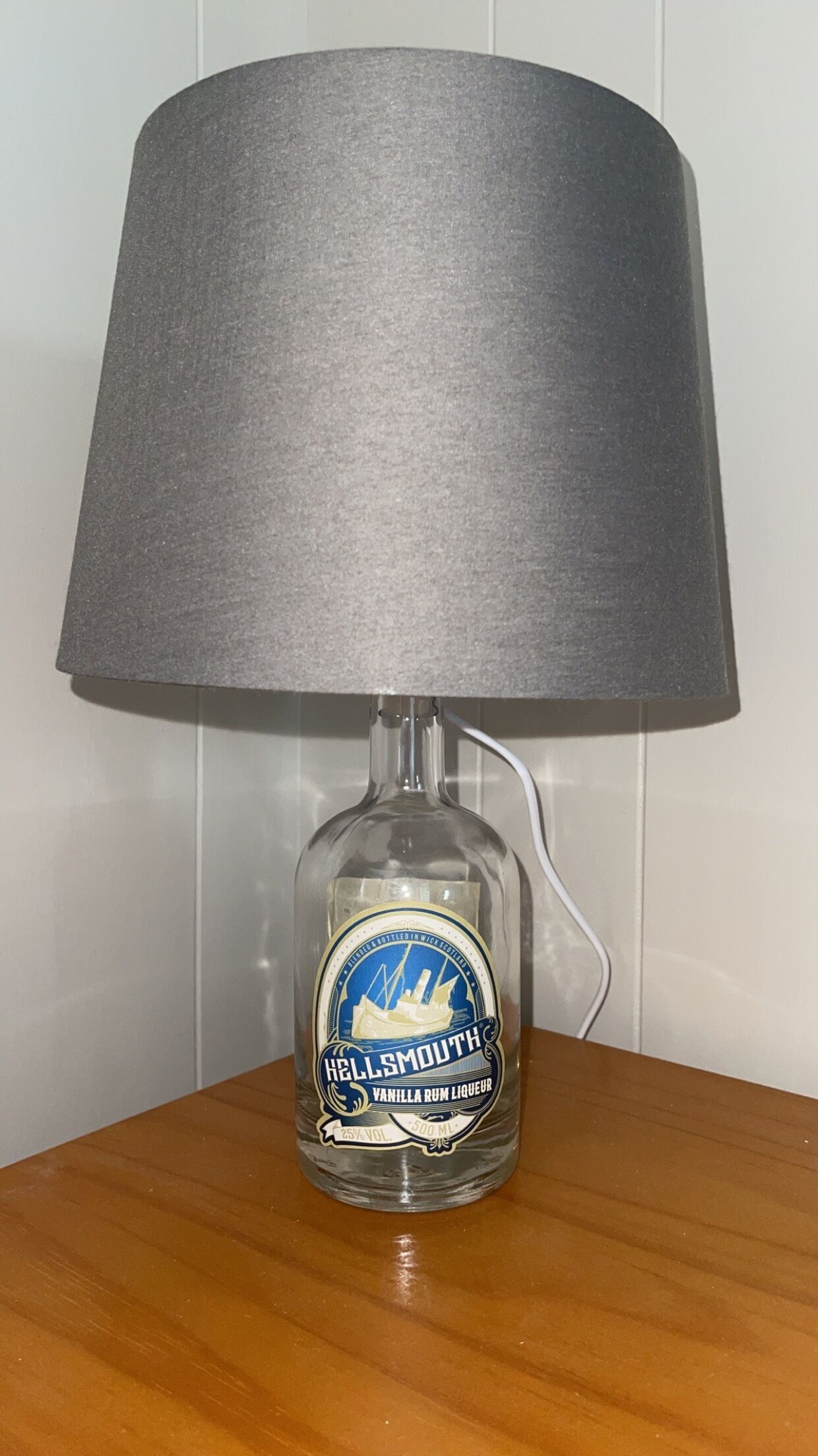A lamp with a clear bottle base, a grey lampshade, and a label reading "HELLSMOUTH VANILLA RUM LIQUEUR 25% VOL 500 ML".