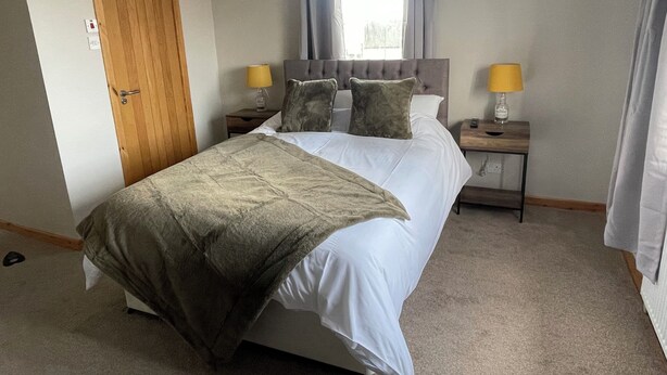 A neatly made bed with a grey blanket, two bedside tables with lamps, a wooden door, and a window with curtains. - Broadhaven Road (Wick)