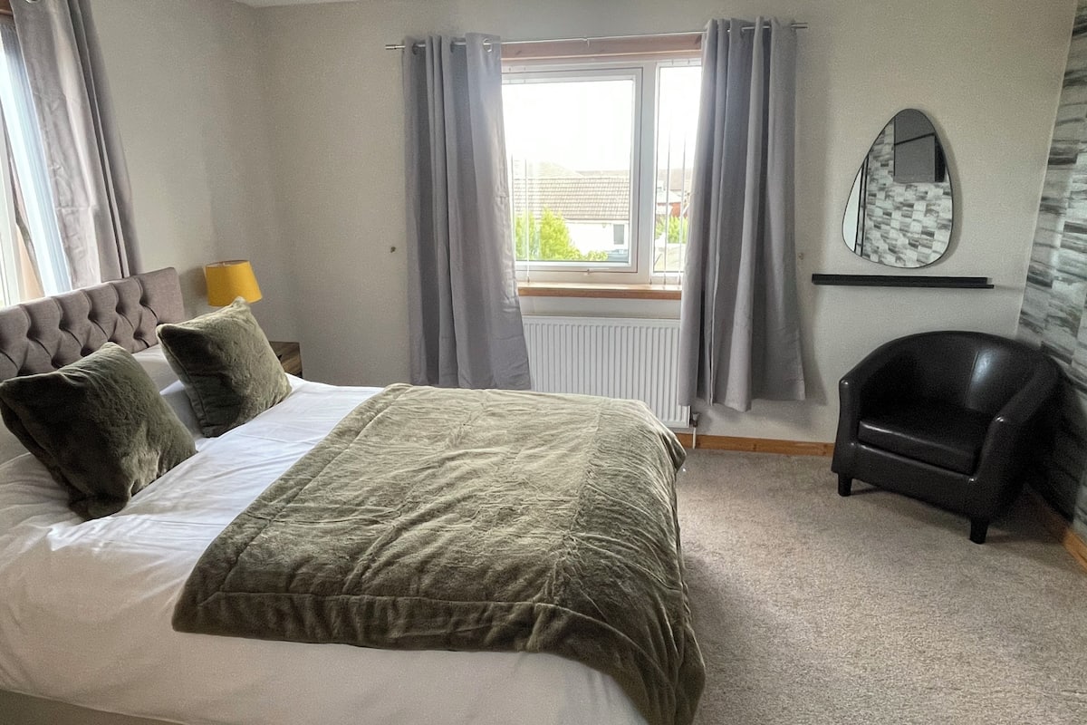 A bedroom with a bed, grey curtains, a window, a lamp, a chair, and a mirror.