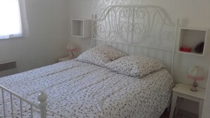 Une chambre avec un lit blanc, une literie à motifs floraux, une tête de lit décorative, des tables de chevet avec des lampes et une petite étagère ornée d’objets décoratifs.