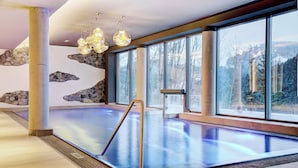 A modern indoor swimming pool with large windows, stone wall art, and a contemporary light fixture. - Königswieser Hof (Koenigswiesen)