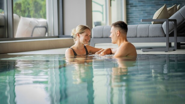 A couple in a swimming pool with a modern interior, including a sofa and a view of greenery outside. - Königswieser Hof (Koenigswiesen)