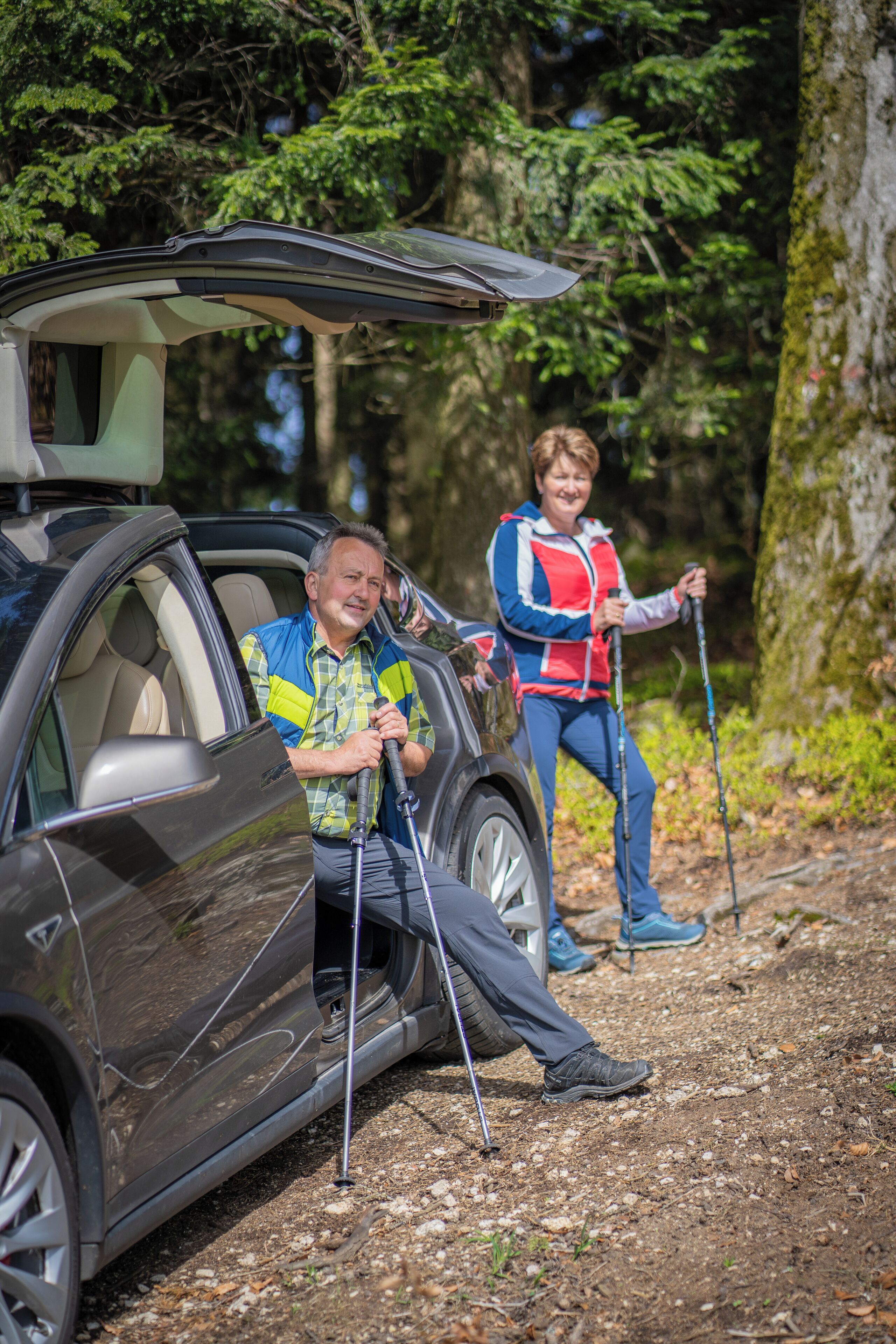 A car parked in a forested area with a person standing next to it holding hiking poles.