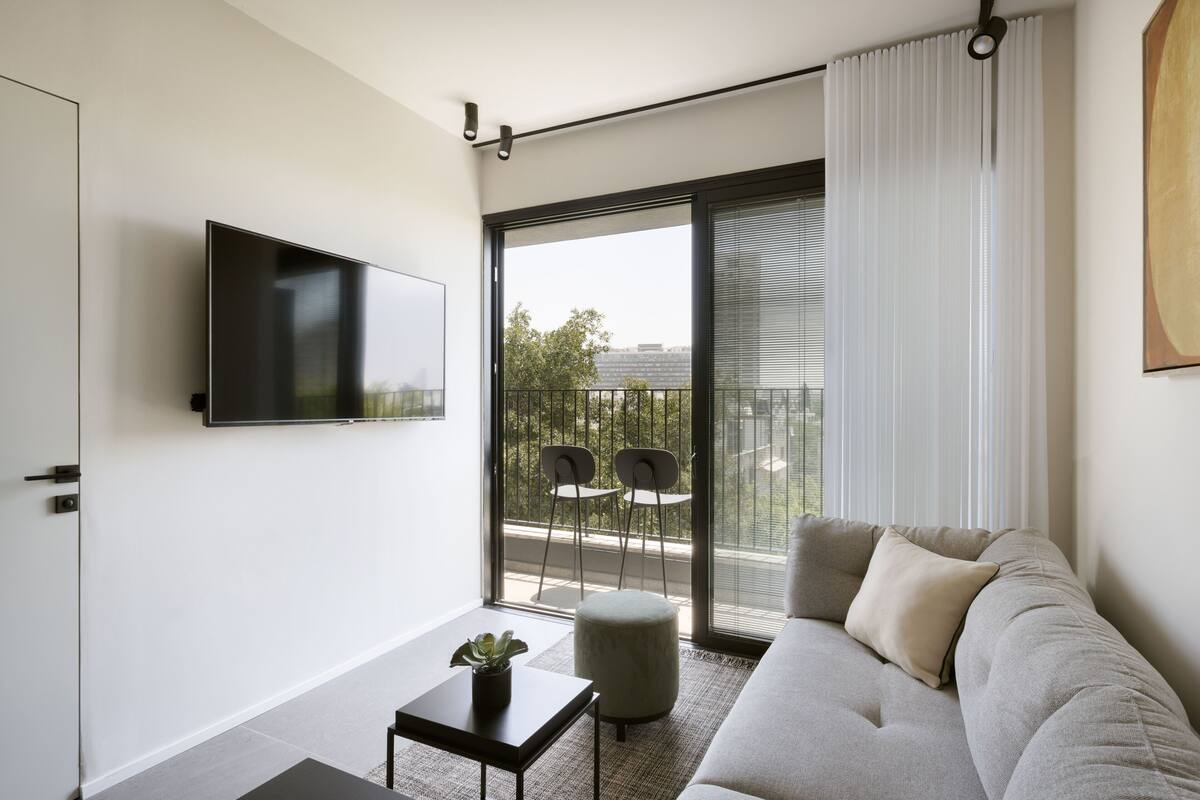 A modern living room with a flat-screen TV mounted on the wall, a grey sofa with cushions, a small table with a plant, and a balcony with chairs.