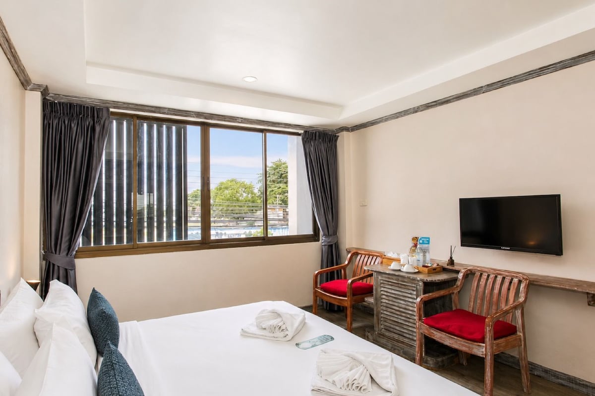 A hotel room with a bed, a desk with chairs, a flat-screen TV, and a large window with curtains.