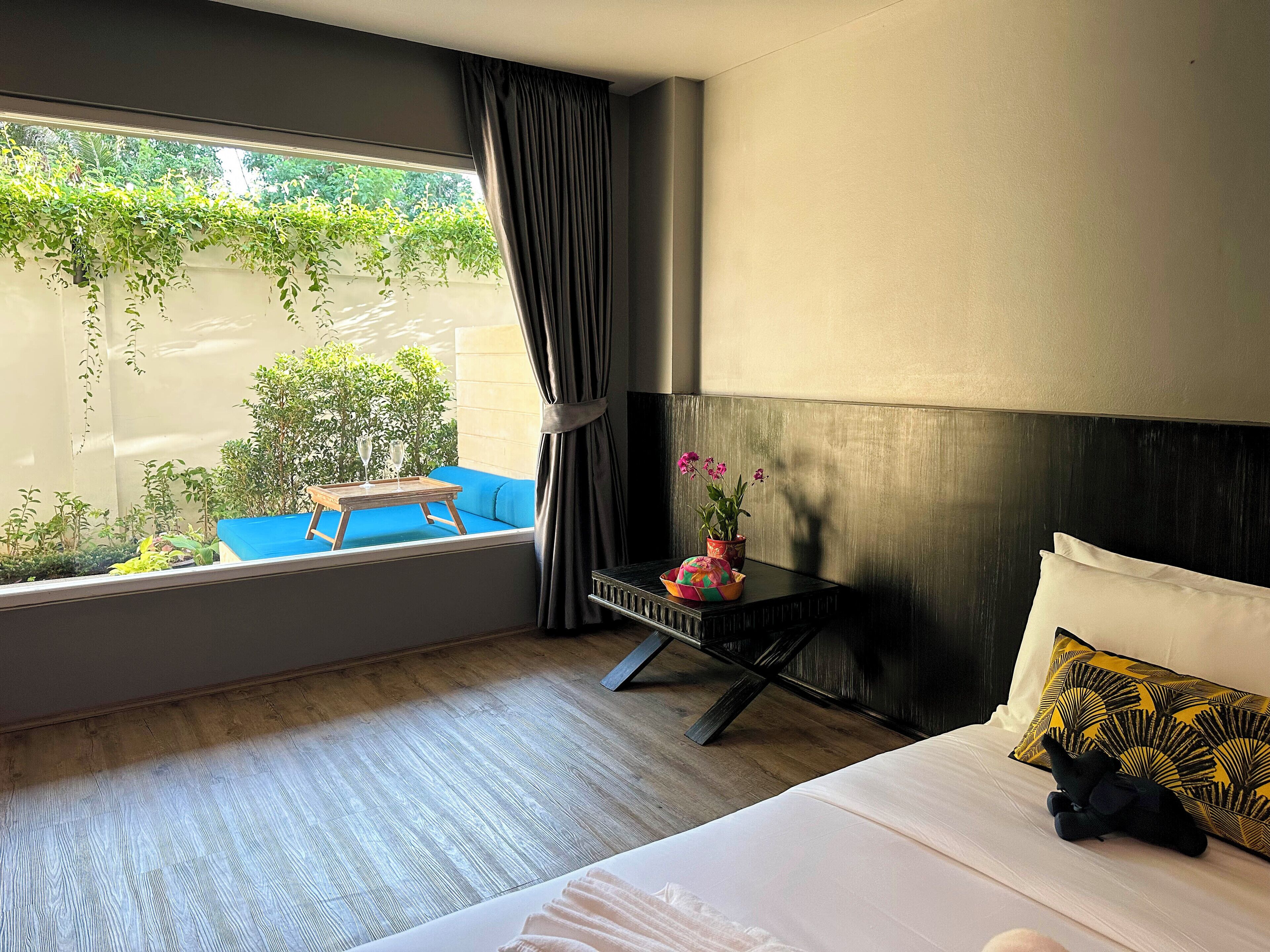 A hotel room with a bed, a small table with a plant and two glasses, a blue chair by the window, and a view of greenery outside.