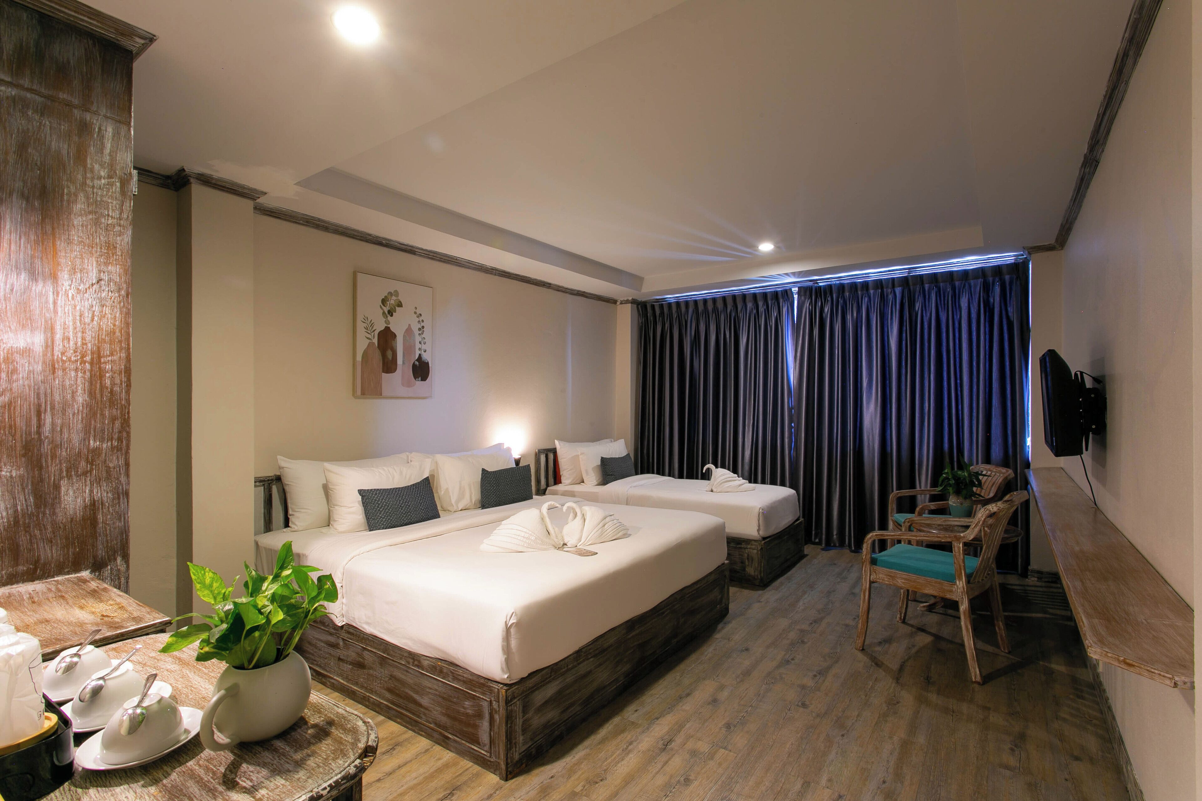 A hotel room with two beds, a wooden floor, a dining table with a plant, and a TV mounted on the wall.