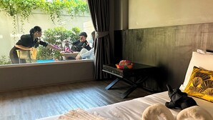 A hotel room with a bed, a window with curtains, a bedside table with a basket, and a small elephant plush toy on the bed. - JALMIN Samui Hotel (Koh Samui)